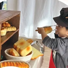 パン屋さんの帽子とエプロンを身につけて、おもちゃのパンを並べる子ども