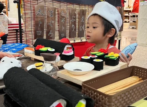 お寿司屋さんの帽子をかぶって、おもちゃのお寿司を並べる子ども